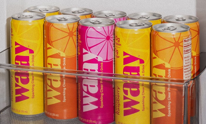Twelve cans of Waay sparkling clear protein drink, in Grapefruit (pink), Lemon Raspberry (yellow), and Orange (orange) flavors, neatly arranged in a refrigerator drawer.